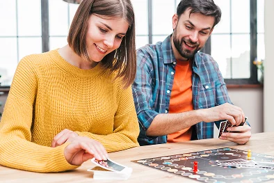 Egy sárga pulóvert viselő, mosolygós nő és egy kockás ingben lévő férfi élvezettel társasjátékot játszanak, ami vidám és barátságos hangulatot teremt.